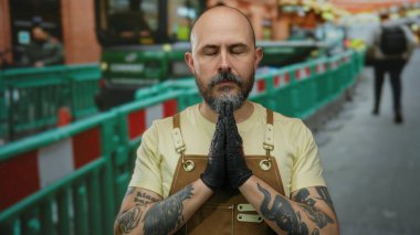 Dövmeli ve sakallı kel adam inşaat alanında gözleri kapalı eldiven ve tulum giyiyor..