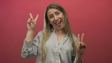 Sarışın, orta yaşlı, gülümseyen bir kadın stüdyoda kırmızı duvarlı barış işareti hareketi gösteriyor. İyimserlik neşe verici..