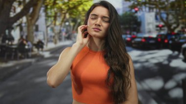 Turuncu tişörtlü kadın, sokakta diş ağrısı için elini yanağına koyup başını arkaya yaslıyor. Fiziksel rahatsızlık..