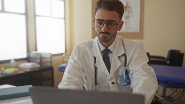 Klinikteki danışma odasında steteskoplu bir adam dizüstü bilgisayardaki verileri inceliyor..