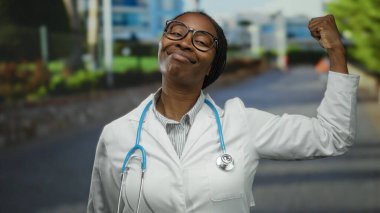 Gözlüklü ve steteskoplu kendine güvenen kadın doktor şehir merkezinde kollarını esnetiyor ve gururlu bir gülümsemeyle şehir ortamında güç ve profesyonellik öneriyor.
