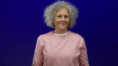 Mavi arka planda gülümseyen bir kadın, kıvırcık gri saçlarını ve pembe kazağını sergiliyor, izole edilmiş bir stüdyoda huzurlu ve mutlu bir yaşlı varlığını yansıtıyor.