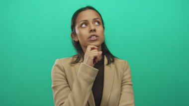 Bronz ceketli bir kadın elini çenesinin üzerine koyup yeşil bir arkaplanda düşünceli ve soyutlanmış bir ifade sergiliyor..