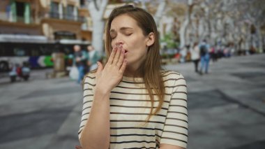 Çizgili bluzlu kadın gözleri kapalı, ağzı kapalı, kalabalık şehir caddesinde esniyor ağaçlarla ve geçen otobüslerle dolu, bitkinlik.