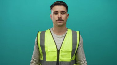 Yansıtıcı yelek giyen genç İspanyol adam, izole edilmiş yeşil duvar arka planına karşı duruyor. Düşünceli ve sakin görünüyor..