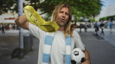 Sarışın ve uzun saçlı bir adam köpük parmağını yukarı doğru tutar ve bir futbol topunu kavrar; ham tutku..