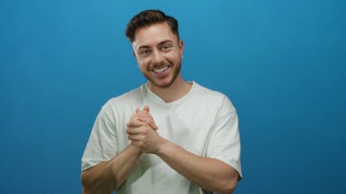 Sakallı genç adam sıcak bir şekilde gülümsüyor, parlak mavi bir arka plan karşısında beyaz gömlek giyiyor, dostça, yaklaşılabilir bir tavır sergiliyor, kurumsal veya yaşam tarzı için mükemmel..