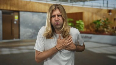 Boş bir şehir caddesinde ahşap bir mağazanın önünde dikilirken göğsünde uzun sarı saçlı bir adam acı çekiyor..