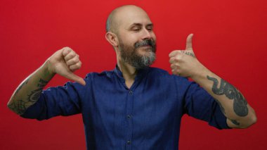 Sakallı kel bir adam, kırmızı arka planda onaylanmış ve onaylanmamış jestler yapıyor, mavi gömlek giyiyor, dövmeler yaptırıyor, yüz ifadelerinde karşıtlık tasvir ediyor..