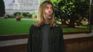 Sarışın adam dudaklarını büzer ve başını hafifçe eğerek yeşil çimenli ön cephe inşa eder..