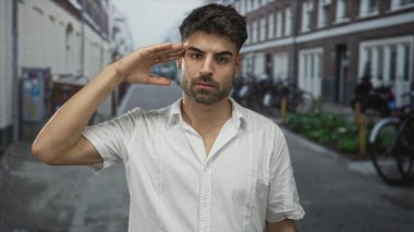 Latin kökenli genç adam, sokağın köşesinde, yumuşak güneş ışığı altında gündelik selamlama jesti ile elini kaldırır; sıcak selamlaşma.