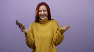 Pürüzsüz mor bir arka planda neşeli bir ifadeyle akıllı telefonu tutan kadın rahat sarı bir kazak giyiyor. Mutluluğu ve modern teknolojiyi temsil ediyor..