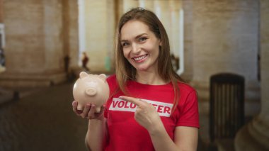 Domuz kumbarası tutan kadın sokakta gülümserken parmağıyla işaret ediyor..