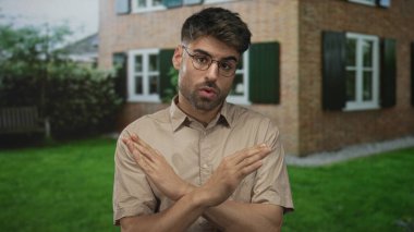 Güneşli bir günde yeşil panjurların altındaki tuğla bina girişinde kollarını kavuşturan gözlüklü bir adam..