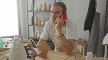 Çanak çömlek atölyesinde akıllı telefon tutan ve defter okuyan adam; konsantrasyon verimliliği planlama.