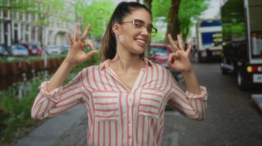 Genç kadın, arabaların ve yapraklı ağaçların olduğu hareketli şehir sokağında el hareketleriyle iyi bir jest yapıyor..