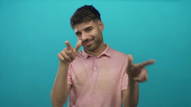 İspanyol adam iki işaret parmağını da stüdyoda yüzsüz bir sırıtışla deniz mavisi duvarına karşı gösteriyor..