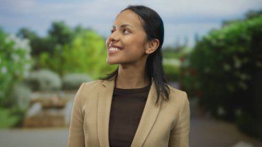 Canlı bir park ortamında gülümseyen bir kadın, açık bir gökyüzünün altında yemyeşil bir hava ile çevrili neşeli bir yüz ifadesi sergiliyor..