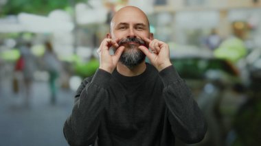 Şehir sokağında dikilen sakallı kel adam kapalı gözlerle ve bulanık şehir geçmişine karşı gevşek tavırlarla memnun görünüyor.