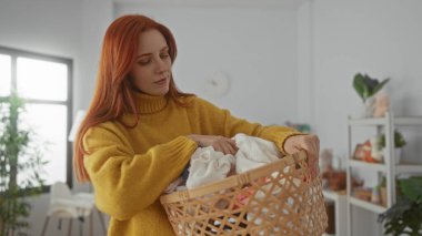 Çamaşır sepetini evdeki rahat çamaşır odasında tutan kadın. Pencereden süzülen güneş ışığı ve sakin atmosferi yükselten dekoratif bitkiler..