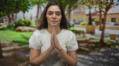 Yeşil ağaçların altında ve halka açık bir parkta çimlerin üzerinde ellerini birbirine bastırarak dua eden bir kadın..