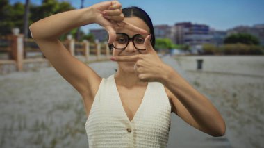 Genç İspanyol kadın dışarıda yeşil bir parkta parmaklarıyla yüzünü çerçeveliyor, doğada yaratıcılığını parlak bir gülümseme ve gözlüklerle ifade ediyor..