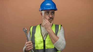 Yansıtıcı yelek giyen ve kasketli genç İspanyol bir adam ingiliz anahtarını düşünceli bir şekilde izole edilmiş kahverengi bir arka plana karşı tutuyor..