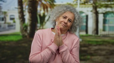 Gri saçlı yaşlı bir kadın, ağaçların altında pembe bir kazak giyerken parkta dua ediyor. Yüzünde huzur ve sükunet ifadesi var..