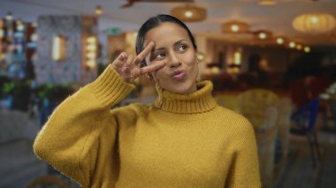 Sarı kazaklı genç İspanyol kadın sıcak bir otelde neşeli ve dost canlısı bir atmosfer sergileyerek neşeli bir barış işareti yapıyor..