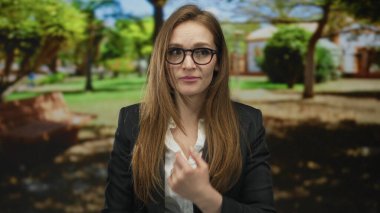 Gözlüklü takım elbiseli kadın yeşil bir parkta tedirgin görünürken göğsüne dokunuyor; belirsizlik kaygısı.