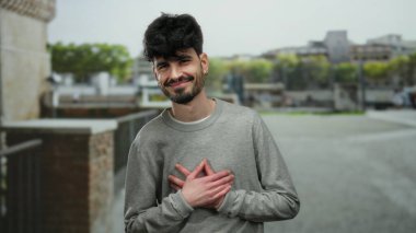 Sokak ortamında göğsünde elleri göğsünde sıcacık gülümseyen genç bir adam..