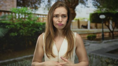 Beyaz elbiseli genç kadın güneş ışığında kollarını kaldırmış yanaklarına parmak uçlarıyla dokunuyor; endişe yansıması.