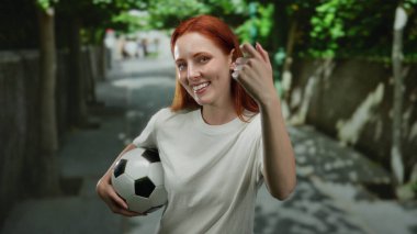 Şehir caddesinde dikilmiş futbol topu tutan bir kadın genç kızıl saçlı güzelliğe odaklanmış canlı şehir manzarasını yakalıyor..