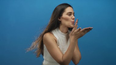 Uzun siyah saçlı, beyaz kolsuz bluz giyen bir kadın mavi stüdyoda kameraya elle öpücük yolluyor..