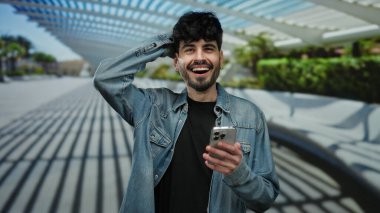 Açık havada elinde akıllı telefonla gezen ve gülen modern mimari geçmişi olan genç bir adam..