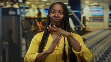 Saçları örgülü, jest yapan, spor salonunda sarı gömlek giyen arka planda bulanık spor aletleri olan bir kadın..