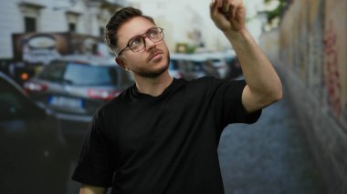 Sakallı genç adam öğleden sonra kalabalık şehir caddesinde olumlu bir tavır sergiliyor ve park halindeki arabalar ve kentsel çevre arasında günlük bir tarz sergiliyor..