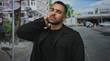 Siyah ceketli adam bulutlu gökyüzü altında şehir binalarının ortasında park halindeki arabaların yanında boynunu tutuyor;.