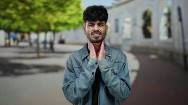 Acılı bir ifadeye sahip genç bir adam, şehir mimarisi ve yeşil ağaçların arasında kot ceket giyip, bir şehir caddesinde boynuna dokunuyor..