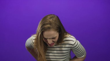 Çizgili üst karın kasları olan beyaz bir kadın mor stüdyoda acı dolu bir ifadeyle öne eğiliyor..