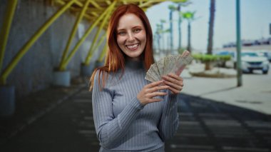 Elinde İsveç banknotları tutan bir kadın, şehir sokaklarında gülümsüyor, kızıl saçlı bir yetişkinin neşeli ifadesini sergiliyor..