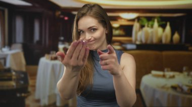 Kadın, restoranın içindeki bulanık masalar ve dekorun ortasında işaret parmağıyla izleyiciye işaret ediyor; davet beklentisi.