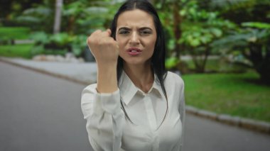 Beyaz gömlekli kadın yemyeşil bir parkta güçlü duygular ifade ediyor. Açık havada güçlü bir öfke ve kararlılık sergiliyor..