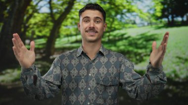 Güneşli bir günde, yeşil ağaçlar ve gölgelerle çevrili bir parkta el kol hareketi yapan genç bir İspanyol, desenli bir gömlek giymiş, elleri açık havada dışa dönük poz veriyor..