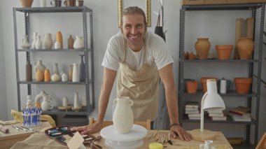 Ahşap masaya yaslanmış önlük giymiş bir adam seramik kavanozlarla dolu bir çömlek atölyesindeki kil vazoya dokunuyor..