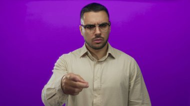 Genç İspanyol gözlüklü adam parlak mor stüdyoyu işaret ediyor; kararlılık odaklı otorite.