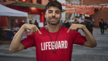 İspanyol adam kırmızı cankurtaran gömleğini işaret ediyor ve kalabalık şehir caddesinde ıslık çalıyor..