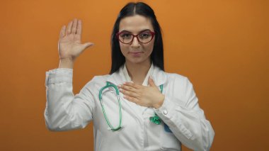 Steteskoplu beyaz önlüklü İspanyol kadın doktor, portakal arkaplanına karşı duruyor ve bağlılığını ve profesyonelliğini gösteren bir taahhüt hareketi yapıyor..