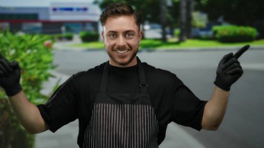 Siyah önlüklü ve eldivenli bir adam şehir sokaklarında çevreyi göstererek gülümsüyor..