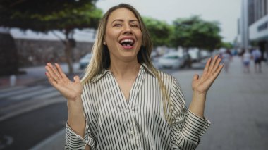 Kadın, arkasında park etmiş arabalar ve tepesinde yapraklı ağaçlar olan yoğun şehir caddesi kaldırımında el kaldırıyor; kendiliğinden mutluluk..